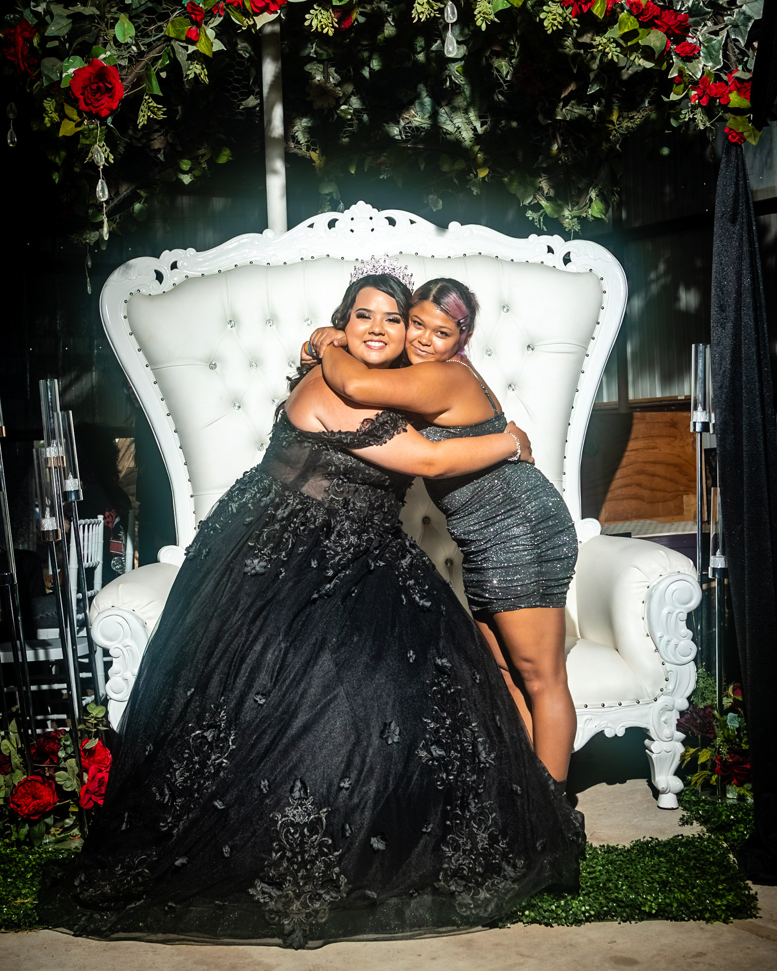 two ladies hugging at an event with a large white chair in the background and flower wreath in the foreground